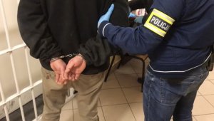 Zatrzymany mężczyzna zakuty w kajdanki na ręce trzymane z tyłu. Obok policjant Wydziału Kryminalnego z opaską z napisem Policja na lewej ręce.