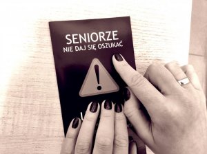 Dłonie kobiety, która wskazuje palcem na ulotce informacyjnej napis: Seniorze. Nie daj się oszukać. Pod napisem znak ostrzegawczy