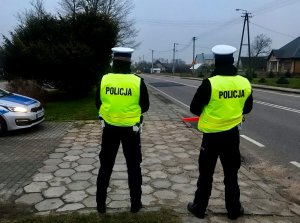 Dwóch policjantów Wydziału Ruchu Drogowego stojących przy ulicy. Po lewej stronie zdjęcia widoczny przód oznakowanego radiowozu. W tle jadący ulicą samochód.