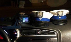Czapki policjantów Wydziału Ruchu Drogowego oraz ręczny miernik prędkości, leżące na podszybiu radiowozu.