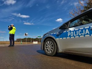 Świeci słońce. Umundurowany policjant stojąc przy ulicy mierzy prędkość pojazdów ręcznym miernikiem prędkości. Po prawej stronie zdjęcia stoi oznakowany radiowóz.