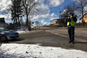 Świeci słońce. W tle droga. Umundurowany policjant stojąc przy jedni mierzy prędkość pojazdów ręcznym miernikiem prędkości. Po lewej stronie zdjęcia stoi oznakowany radiowóz.