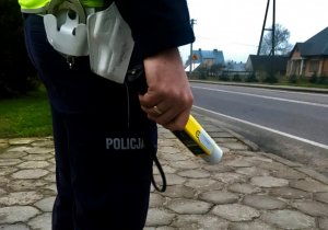 Umundurowany policjant trzyma w dłoni żółty alkomat. W tle droga.