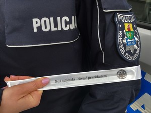 Policjantka trzyma w dłoni opaskę odblaskową z logiem KPP w Kolnie i napisem Noś odblaski - świeć przykładem.